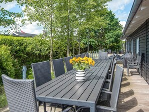 An outdoor patio with a long table and wicker chairs, surrounded by greenery and a building. - 5 Star Holiday Home in Hadsund (Hadsund)
