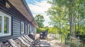A covered outdoor area with sun loungers and a table, surrounded by trees and a fence. - 5 Star Holiday Home in Hadsund (Hadsund)
