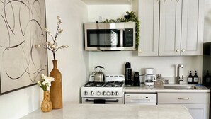 Cocina moderna con armarios blancos, microondas integrado, estufa a gas, cafetera y dos taburetes de mimbre.