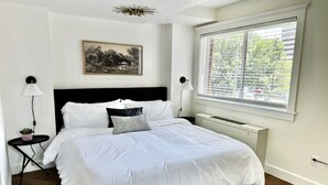 Un dormitorio con cama, radiador, ventana con persianas, un cuadro en la pared y una mesita de noche con lámpara.