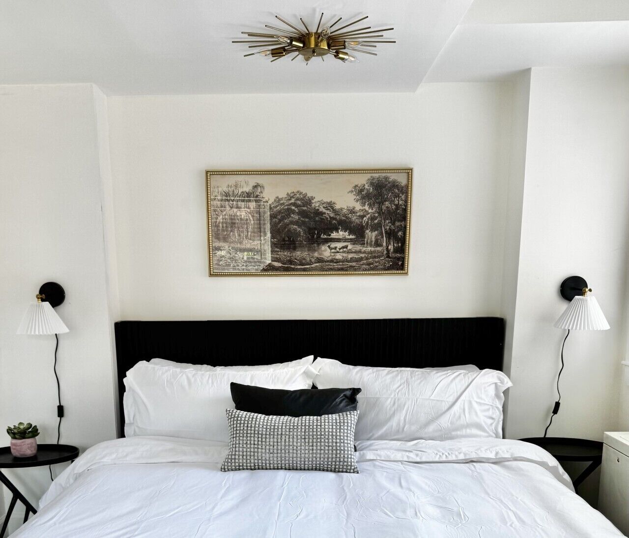A neatly made bed with white linens, a black headboard, and a black and white patterned pillow. There is a framed black and white landscape painting above the bed.