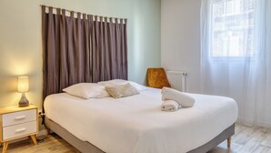 A bedroom with a bed, a nightstand, a lamp, a chair, a window with curtains, and a radiator. - XAVIER – F2 cosy avec balcon à Rouen (Rouen)
