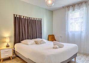 A bedroom with a bed, a nightstand, a lamp, a chair, a window with curtains, and a radiator. - XAVIER – F2 cosy avec balcon à Rouen (Rouen)