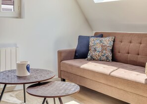 Ein gemütliches Zimmer mit einem braunen Sofa, zwei gemusterten Kissen, einer weißen Tasse und einem kleinen runden Tisch mit einem gewobenen Unterblatt.