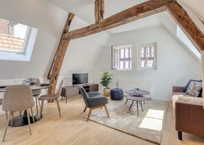 Ein Loft-Zimmer mit freiliegenden Holzbalken, einer Essecke, einer Sitzecke mit Sofa und Sesseln, einem Fernseher und einem Blumentopf.