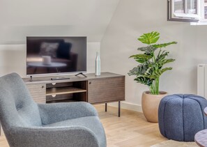 A modern living room with a flat-screen TV, a grey armchair, a blue ottoman, and a large potted plant. - JACKY - Joli 2 pièces avec vue sur l'Eglise Saint Maclou (Rouen)