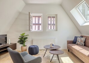 A modern loft bedroom with a sloped ceiling, a large window, a radiator, a sofa, a TV, a plant, and a small round table. - JACKY - Joli 2 pièces avec vue sur l'Eglise Saint Maclou (Rouen)