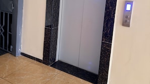 Un ascensor con un panel de control en la pared, una puerta con una grieta visible y un suelo de baldosas.