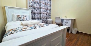 Un letto rifatto con una coperta a motivi, una testiera bianca, una scrivania con una sedia e un cestino.