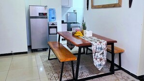 Una cocina pequeña con refrigerador, dispensador de agua y estufa. Hay una mesa con un banco y un mantel estampado.