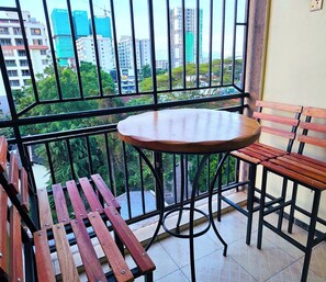 Un balcone con un tavolo rotondo e panche di legno, affaccia su una città con edifici e spazi verdi.