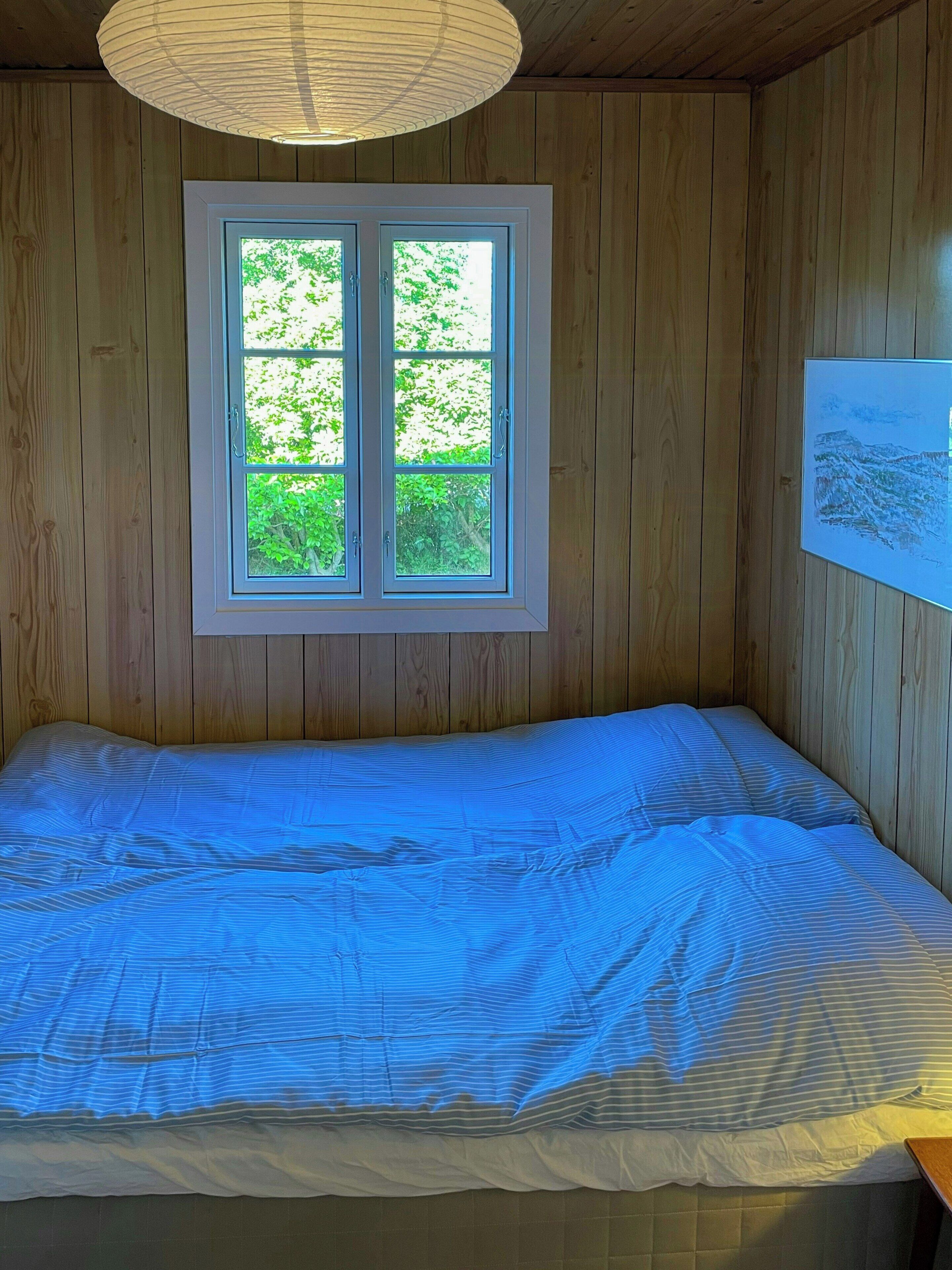 A room with wooden panel walls, a bed with a blue-striped bedspread, a window with a view of greenery, and a framed artwork on the wall.