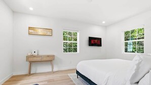 Un dormitorio con cama, escritorio, televisor y dos ventanas con vistas a la vegetación.