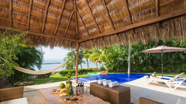 Un espace aménagé au bord de la piscine, doté d’un toit de chaume, de chaises longues, d’une table où sont disposés des fruits et des boissons, et offrant une vue sur la mer.