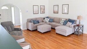 Ein Wohnzimmer mit einem Ecksofa, Poufs, einem grauen Sessel und einem kleinen Beistelltisch.