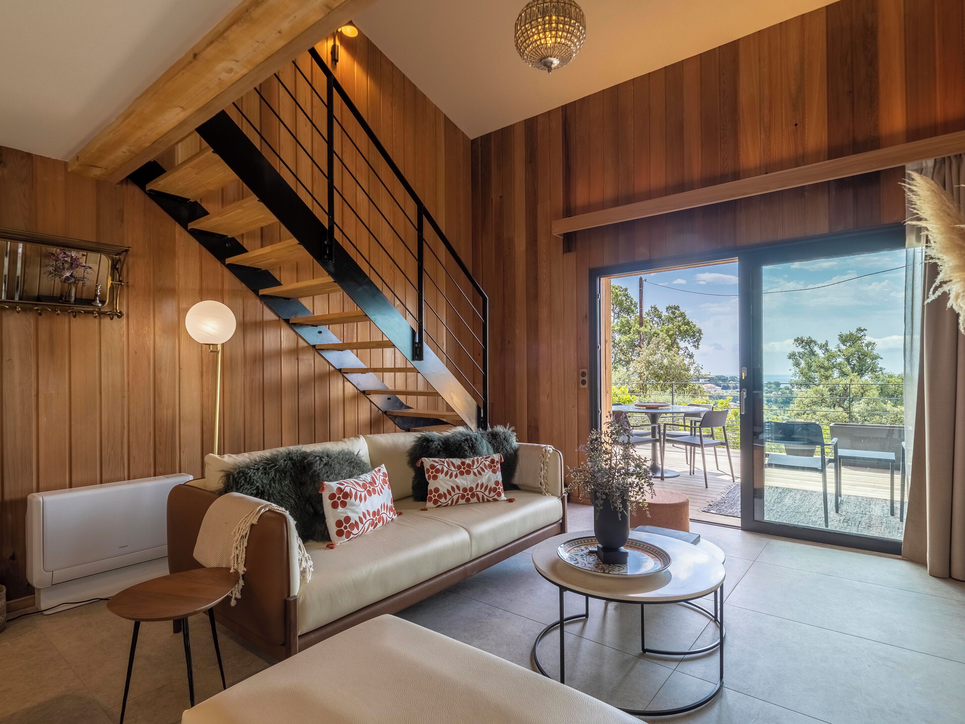 Un salon moderne avec un escalier en bois, un canapé agrémenté de coussins décoratifs, une table basse ronde et une vue à travers une porte vitrée.