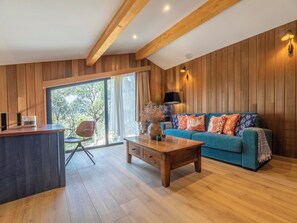 Ein modernes Wohnzimmer mit Holzboden, einem blauen Sofa mit gemusterten Kissen, einem Holztisch und einem großen Fenster mit Vorhängen.