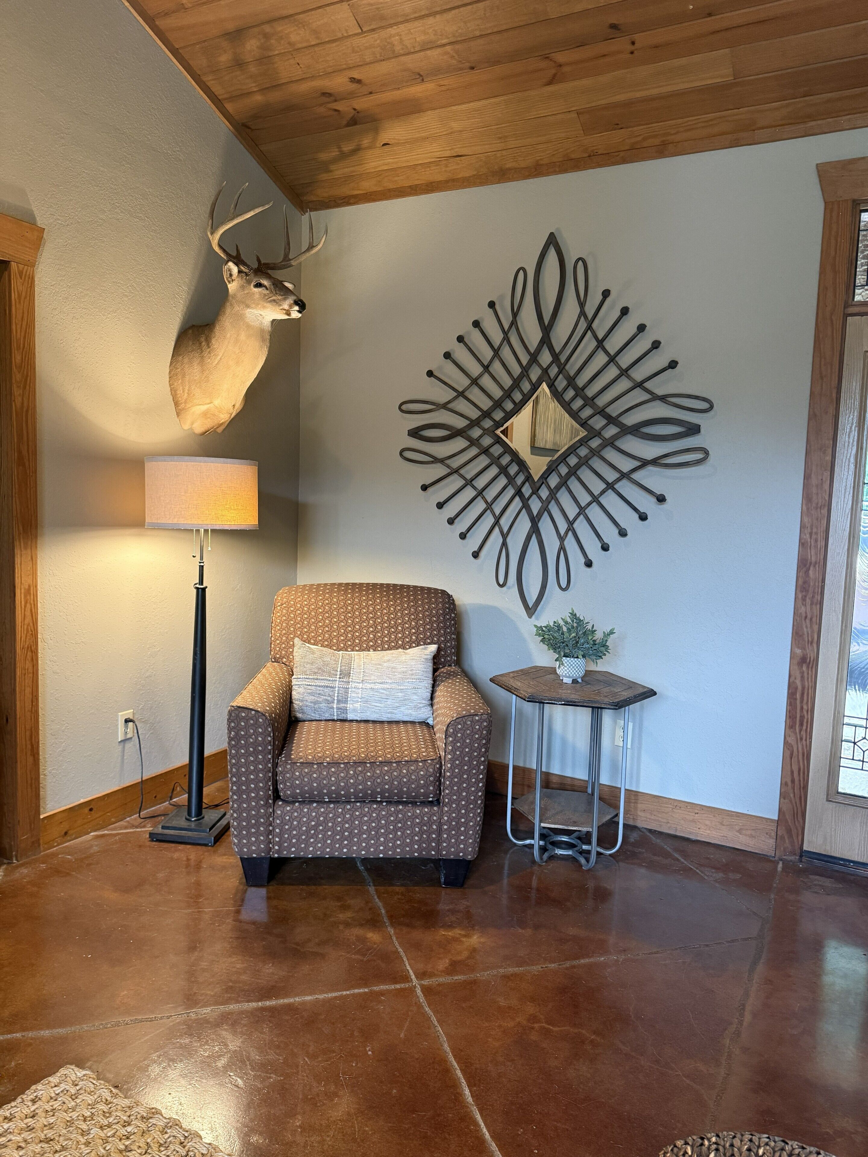 Une pièce comprenant un fauteuil à motifs, une lampe, une tête de cerf fixée au mur et un élément décoratif en métal.