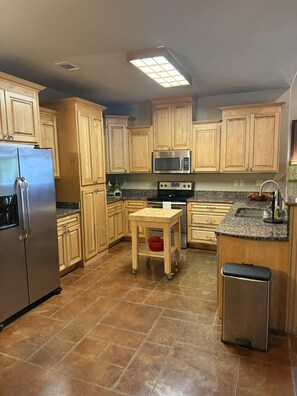 A kitchen with wooden cabinets, a stainless steel refrigerator, a microwave, an oven, a granite countertop, a small table, and a trash bin.