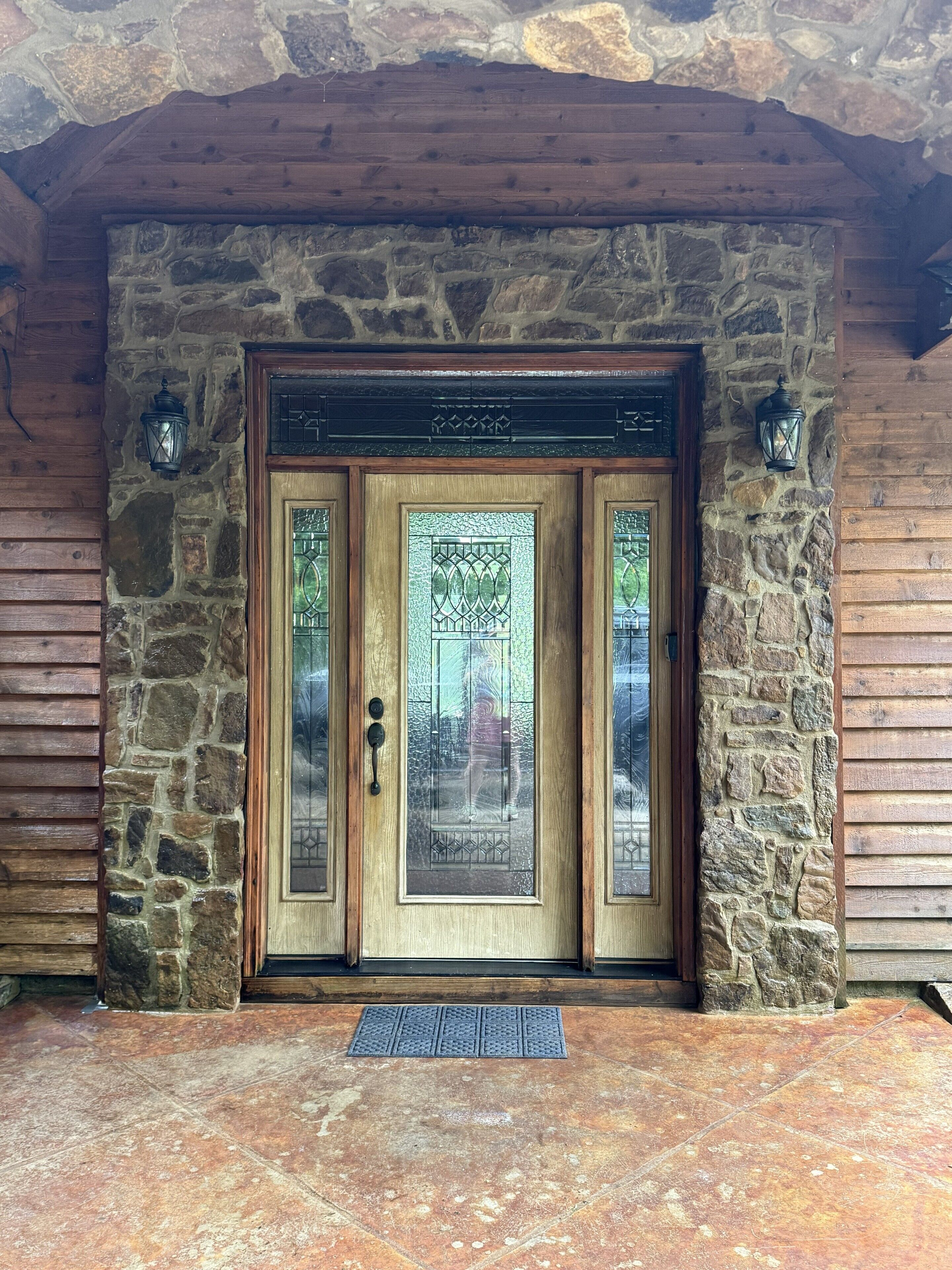 Une entrée en pierre, avec un mur de pierres, surmontée d’une porte en bois ornée de panneaux de vitraux et dotée d’une poignée de porte en métal.
