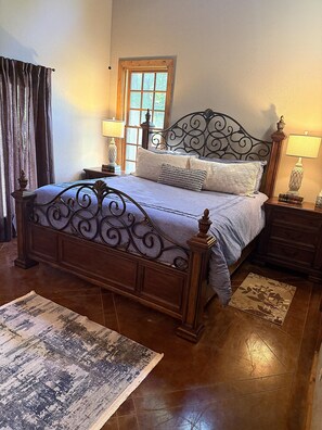 A bedroom with a wooden bed, a window with curtains, a nightstand with a lamp, and a rug on the floor.
