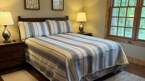 Quarto com cama, duas mesas de cabeceira com abajures, uma janela com vista para árvores e dois painéis decorativos fixados na parede.