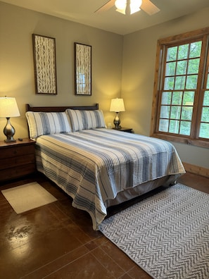 Un dormitorio con cama, dos mesitas de noche con lámparas, una ventana con vistas a árboles y dos paneles decorativos en la pared.