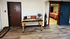 A hotel lobby with a wooden bench, a fire alarm, a sign indicating a restroom, and a bed in the background.