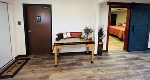Un hall d’hôtel avec un banc en bois, un détecteur d’incendie, un panneau indiquant les toilettes, et un lit en arrière-plan.