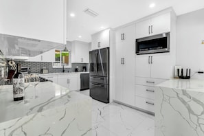 A modern kitchen with white cabinetry, a stainless steel refrigerator, a built-in microwave, and a marble countertop. - NEW Beach Block Villa, Pool, Games (Pompano Beach)
