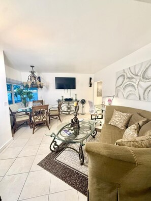 Ein Wohnzimmer mit einer Couch, einem Glastisch, einem Fernseher, einer Chandelieber und einem Essbereich mit Stühlen und einem Tisch.