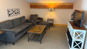A cozy living room with a sofa, ottoman, coffee table, and a TV stand with a flat-screen TV. - Flight Deck - 1-bedroom condo on Beaver Island, your perfect island retreat! (Beaver Island)