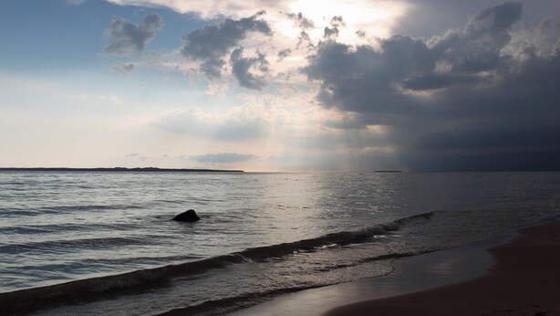 Une plage avec des vagues, une terre lointaine à l’horizon et un ciel nuageux.