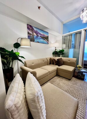 Ein Wohnzimmer mit einem beigefarbenen Ecksofa, zwei Stehlampen, einem großen Gemälde von einer Berglandschaft und einem Fenster mit Vorhängen.