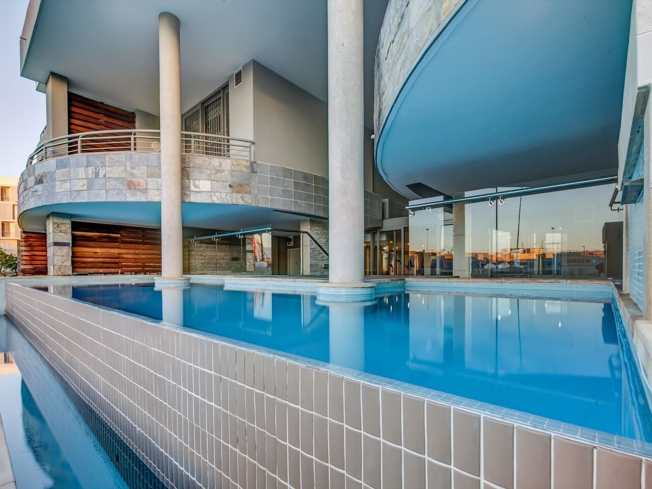 An indoor swimming pool with a tiled edge, a wooden deck area, and a glass balustrade.