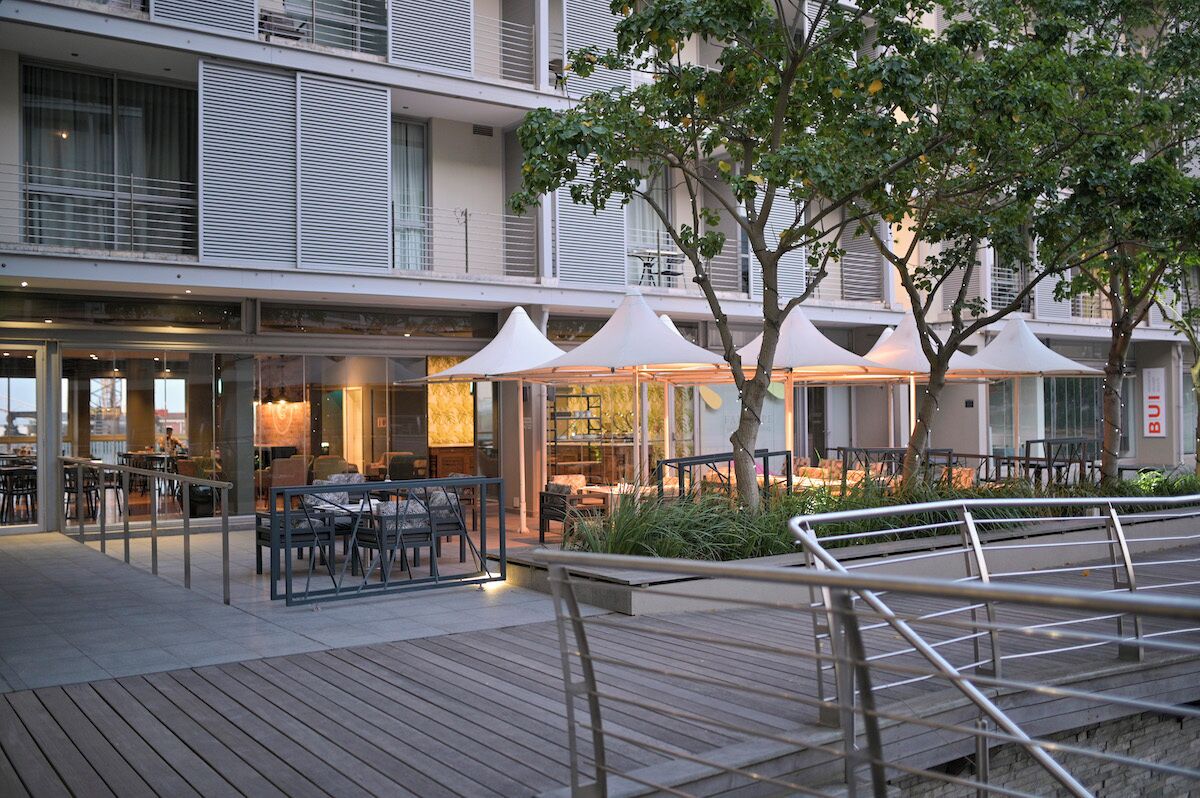 An outdoor dining area with tables and umbrellas, a wooden deck, and a building with large windows and balconies.