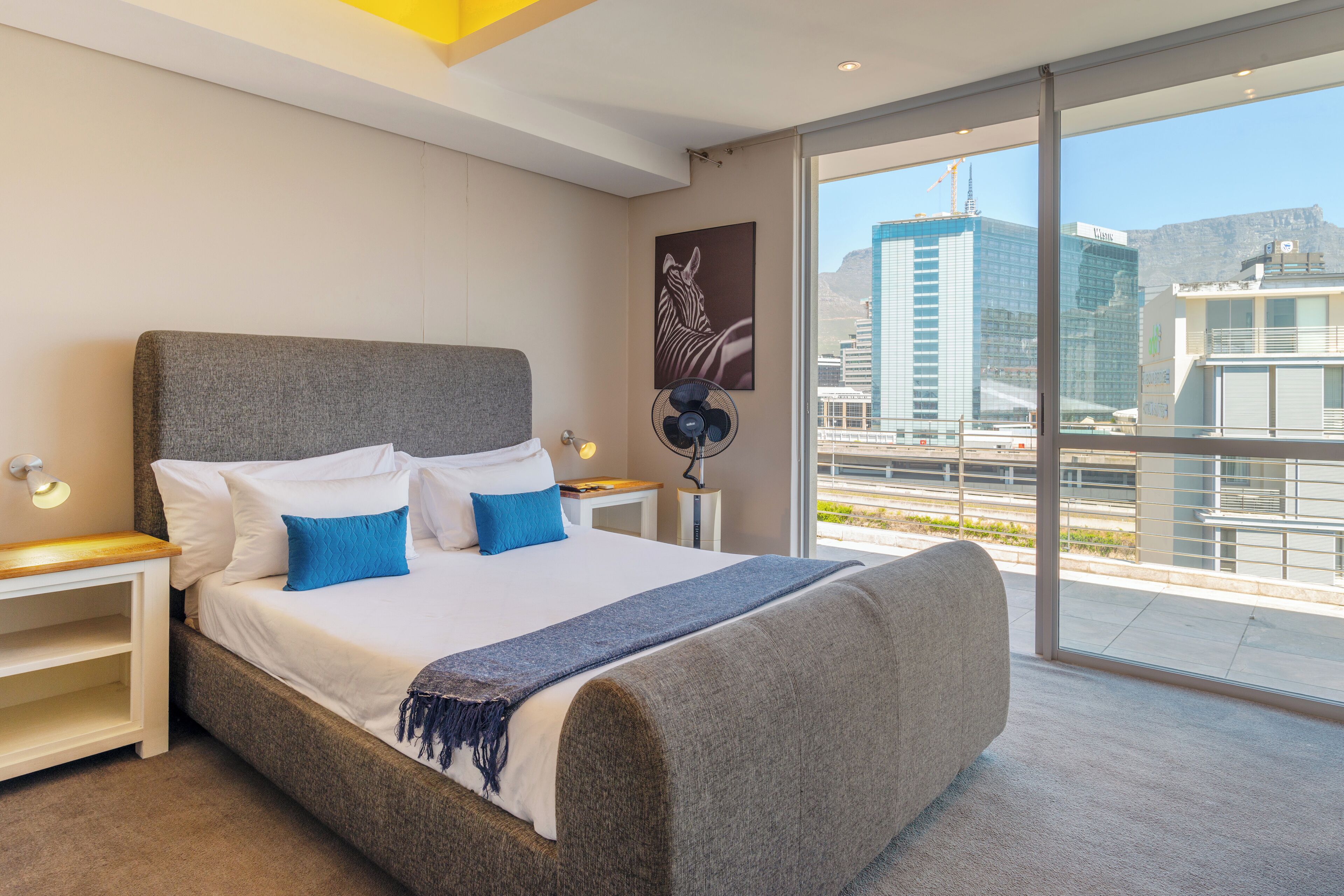 A modern hotel room with a large bed, a bedside table, a floor lamp, and a view of the cityscape.