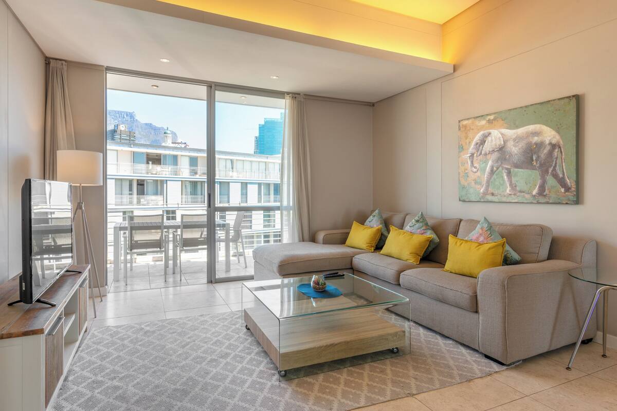 A modern living room with a glass coffee table, a sofa with colorful cushions, a flat-screen TV, and a painting of an elephant on the wall.