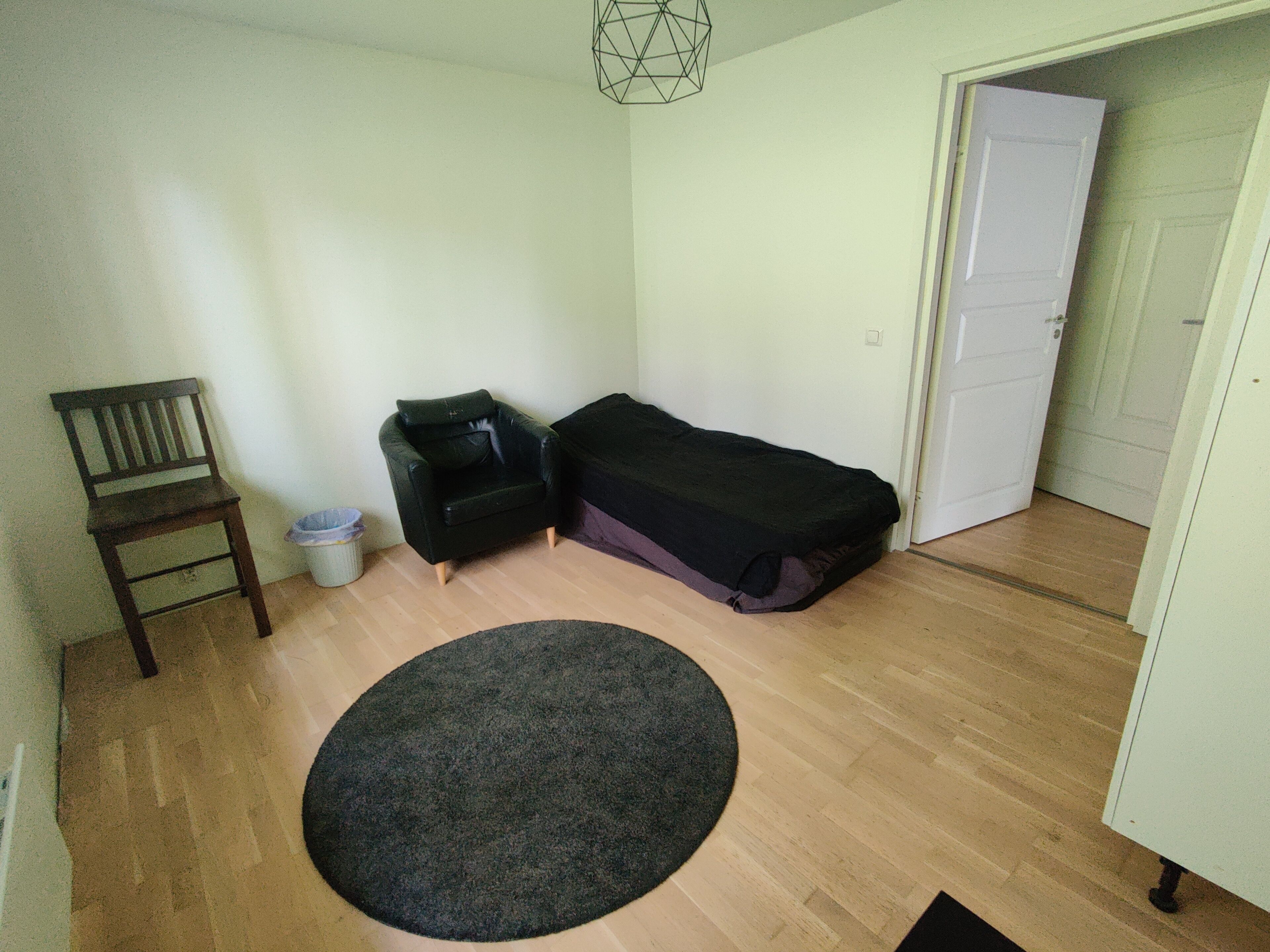 Una habitación pequeña y poco amueblada con piso de madera, una sola silla, un sofá negro, una cama y un tapete redondo.