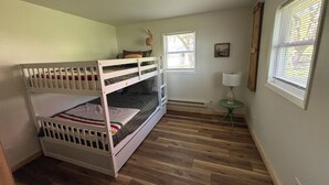 A bunk bed room with a radiator, a small table with a lamp, and two windows with blinds.