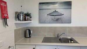 A kitchen with a stainless steel sink, a kettle, and a shelf with various items including a red bag.