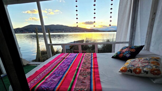 Ein Bett mit bunten Kissen und Blick auf einen See bei Sonnenuntergang.