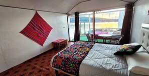 A room with a bed, a table, chairs, a window with a view of a body of water, and a patterned wall hanging. - Uros Titicaca Sunshine (Puno)