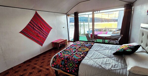 A room with a bed, a table, chairs, a window with a view of a body of water, and a patterned wall hanging.