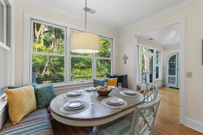 A dining area with a round table set for four, large windows with a view of greenery, a cross on the wall, and a light blue cabinet. - 3BR East Beach Kiawah retreat with marsh and wildlife views! (Kiawah Island)