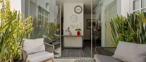Eine sonnendurchflutete Terrasse mit Korbmöbeln, einer Glastür zu einem Innenraum und einer Uhr an der Wand.