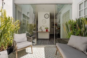 Eine sonnendurchflutete Terrasse mit Korbmöbeln, einer Glastür zu einem Innenraum und einer Uhr an der Wand.