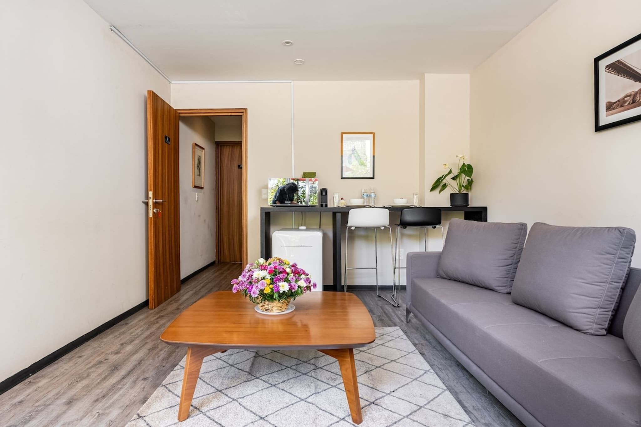 Una habitación moderna con una mesita de centro de madera, un sofá gris, una kitchenette y una puerta que da a otra habitación.