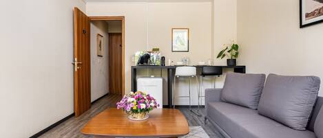 Una habitación moderna con una mesita de centro de madera, un sofá gris, una kitchenette y una puerta que da a otra habitación.