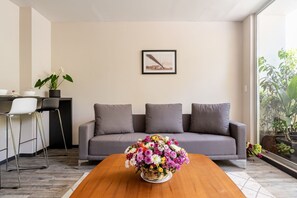 Ein modernes Wohnzimmer mit grauem Sofa, einem Holztisch mit floraler Dekoration, einem gerahmten Bild an der Wand und Blick ins Grüne durch ein großes Fenster.
